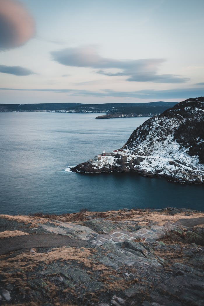 Stony shore near calm water of sea flowing near cliff covered with snow in nature on winter day against cloudy sky